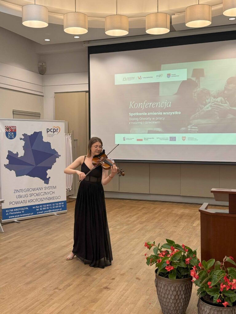 Na zdjęciu widzimy młodą kobietę grającą na skrzypcach, ubraną w elegancką czarną suknię. Stoi na drewnianej podłodze w sali konferencyjnej. W tle znajduje się ekran projekcyjny, na którym wyświetlony jest slajd konferencji z hasłem „Konferencja. Spotkanie zmienia wszystko. Dialog Otwarty w pracy z rodziną i dzieckiem” oraz logotypami organizatorów, takimi jak Wielkopolska, PCPR i Powiat Krotoszyński. Obok ekranu ustawiony jest roll-up z mapą powiatu krotoszyńskiego i informacjami kontaktowymi PCPR. Na pierwszym planie, po prawej stronie, znajdują się dwie duże donice z czerwonymi kwiatami. Sufit zdobią okrągłe lampy, dopełniając atmosferę wydarzenia.