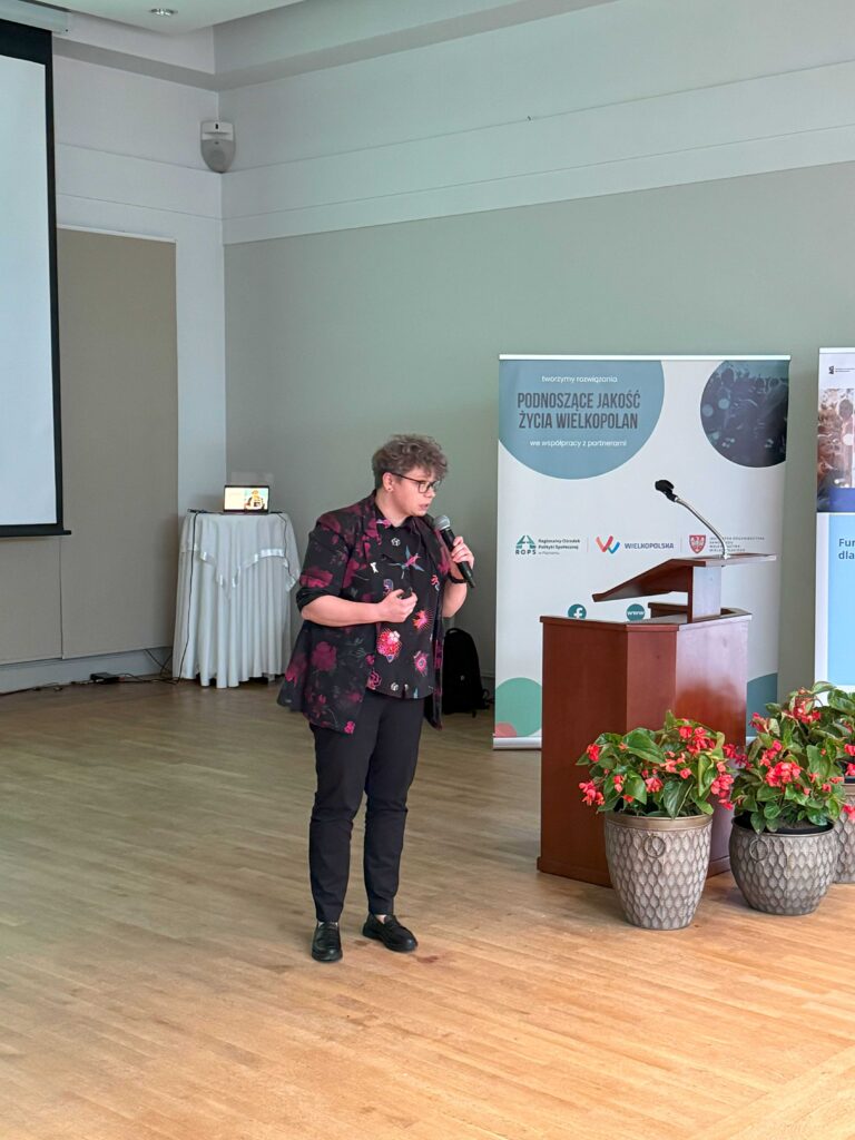 Na zdjęciu znajduje się kobieta przemawiająca na scenie. Osoba trzyma mikrofon i stoi obok drewnianej mównicy. W tle widoczne są dwa roll-upy z napisami, z których jeden zawiera tekst: „tworzymy rozwiązania PODNOSZĄCE JAKOŚĆ ŻYCIA WIELKOPOLAN we współpracy z partnerami”. Na scenie znajdują się również trzy donice z czerwonymi kwiatami. Pomieszczenie ma drewnianą podłogę i jasne ściany.