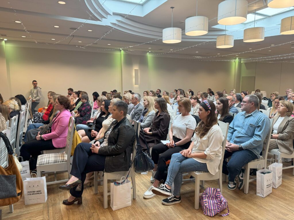 Na zdjęciu widzimy grupę osób siedzących na krzesłach w przestronnej sali konferencyjnej. Na podłodze znajdują się torby z napisem „PODNOSIMY JAKOŚĆ ŻYCIA WIELKOPOLAN”, co sugeruje, że wydarzenie ma związek z inicjatywami społecznymi lub edukacyjnymi. W tle można dostrzec sufit z nowoczesnym oświetleniem i dużymi lampami wiszącymi, co nadaje miejscu elegancki i profesjonalny charakter. Uczestnicy konferencji są ubrani w różnorodne stroje, od swobodnych po bardziej formalne, co wskazuje na otwarty charakter spotkania. Atmosfera sugeruje, że osoby te słuchają prelegenta lub uczestniczą w prezentacji.