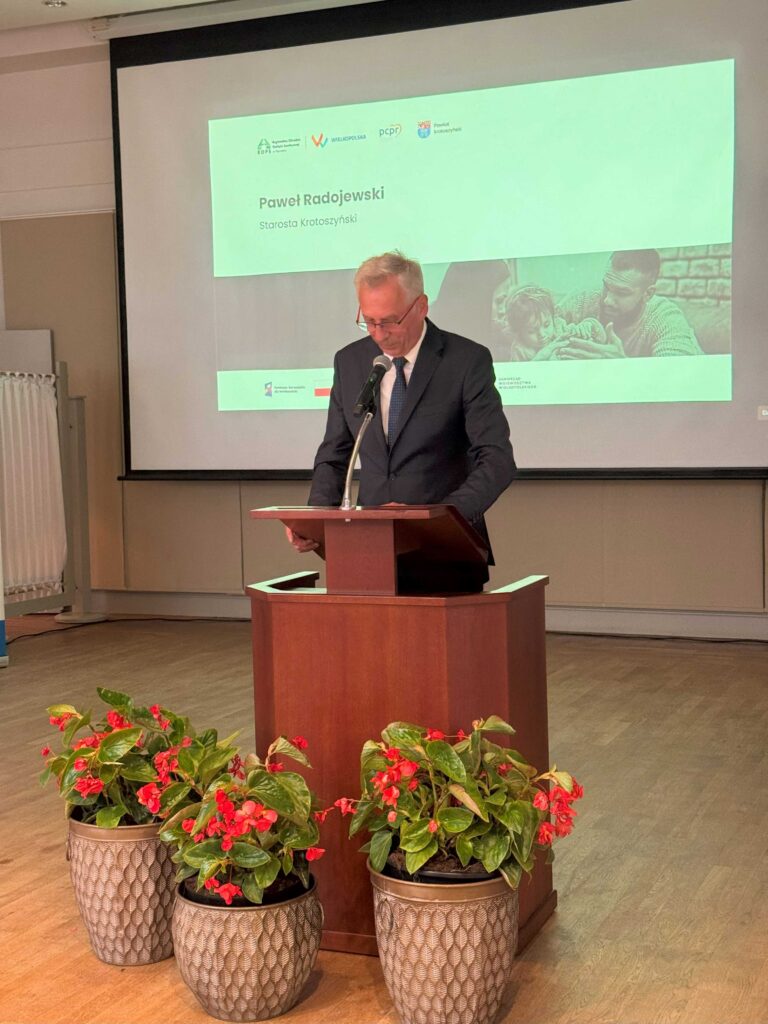 Zdjęcie przedstawia mężczyznę stojącego za drewnianą mównicą, ubranego w garnitur i krawat. Przed mównicą ustawione są trzy doniczki z czerwonymi kwiatami, które dodają elegancji przestrzeni. W tle widoczny jest ekran projekcyjny, na którym wyświetlona jest prezentacja. Na ekranie znajduje się tekst: „Paweł Radojewski Starosta Krotoszyński” oraz logotypy różnych instytucji, takich jak Powiat Krotoszyński, PCPR i Wielkopolska. Dodatkowo w prezentacji widoczny jest obraz przedstawiający dorosłego mężczyznę trzymającego dziecko.
