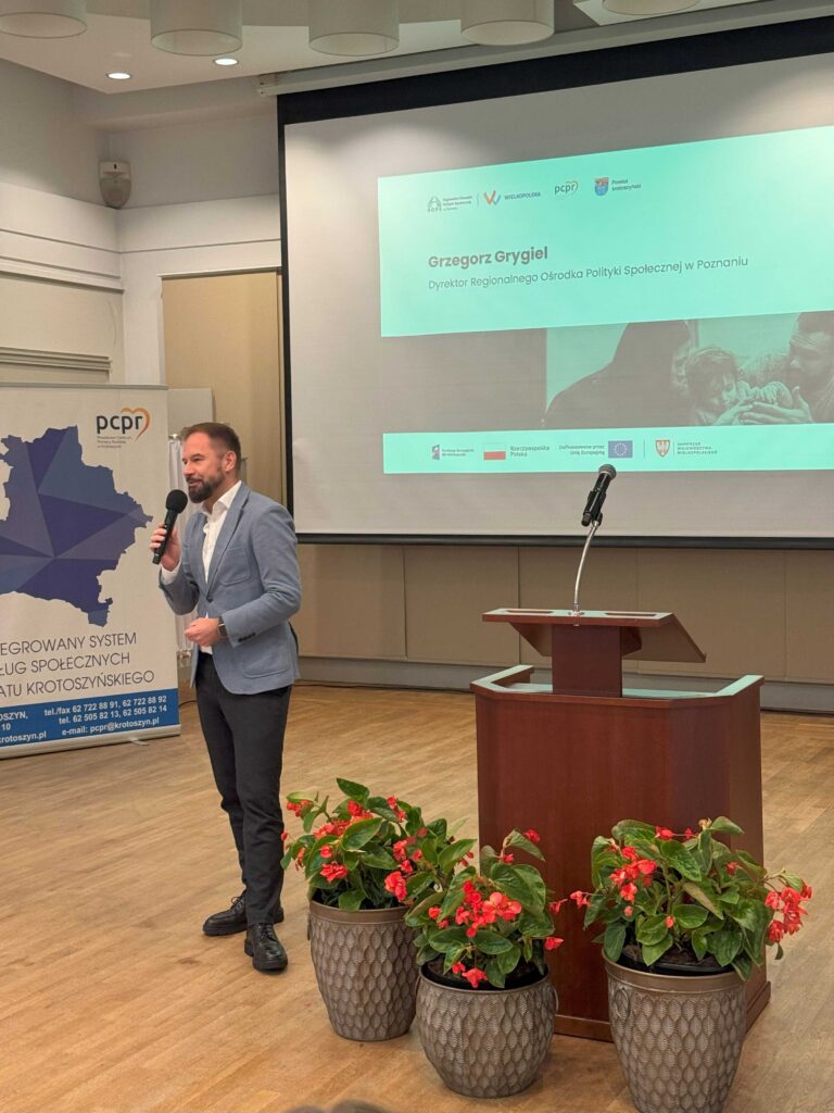 Na zdjęciu znajduje się mężczyzna trzymający mikrofon, stojący obok mównicy w sali konferencyjnej. Na ekranie za nim wyświetlana jest prezentacja z napisem: „Grzegorz Grygiel, Dyrektor Regionalnego Ośrodka Polityki Społecznej w Poznaniu”. W tle widać również baner z napisem „ZINTEGROWANY SYSTEM USŁUG SPOŁECZNYCH POWIATU KROTOSZYŃSKIEGO” oraz dane kontaktowe. Na pierwszym planie znajdują się trzy donice z kwiatami, które dodają przestrzeni estetycznego charakteru.