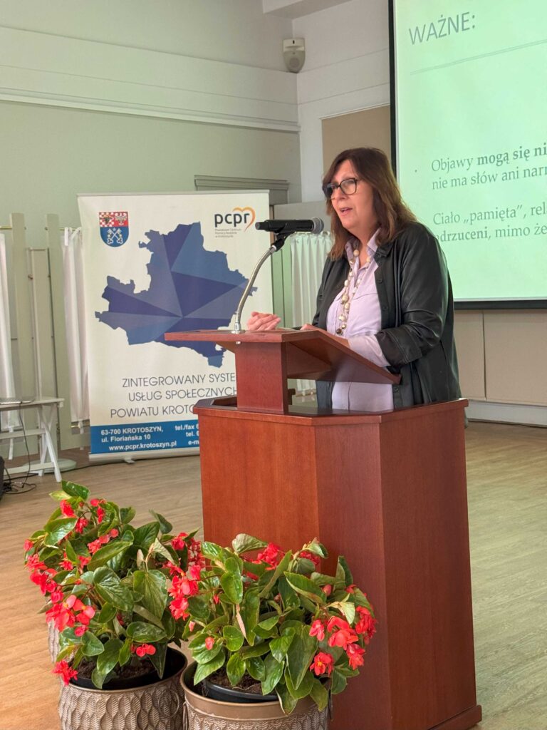 Zdjęcie przedstawia osobę stojącą przy mównicy, która wygłasza prezentację. W tle widoczne są dwa banery. Jeden zawiera mapę Polski z zaznaczonym powiatem krotoszyńskim oraz logo Powiatowego Centrum Pomocy Rodzinie (PCPR). Drugi baner prezentuje nazwę inicjatywy „Zintegrowany System Usług Społecznych Powiatu Krotoszyńskiego” wraz z danymi kontaktowymi instytucji. Na ekranie obok mównicy widnieje fragment prezentacji z nagłówkiem „WAŻNE:” i tekstem dotyczącym pamięci ciała oraz relacji międzyludzkich. Na pierwszym planie znajdują się trzy doniczki z czerwonymi kwiatami, które nadają przestrzeni elegancki charakter.