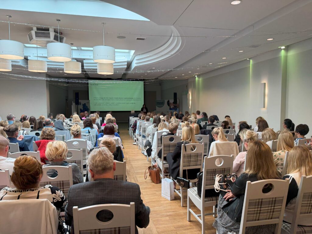 Zdjęcie przedstawia dużą grupę ludzi siedzących na krzesłach w przestronnej sali konferencyjnej. Na pierwszym planie widoczny jest ekran projekcyjny. W tle znajduje się osoba prowadząca prezentację. Sala charakteryzuje się nowoczesnym wystrojem, jasnym oświetleniem i eleganckimi dekoracjami na suficie, tworząc profesjonalną atmosferę wydarzenia.