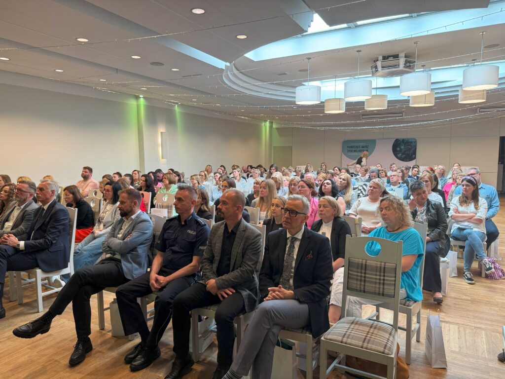 Zdjęcie przedstawia dużą grupę osób siedzących na krzesłach w sali konferencyjnej. W tle widoczne są jasne ściany, nowoczesne oświetlenie sufitowe oraz dekoracje w postaci wiszących światełek. Na ścianie znajduje się tablica z napisem „Pomocniczy Ośrodek Pracy Pedagogicznej”. Wydarzenie wygląda na formalne, a uczestnicy są elegancko ubrani. Na pierwszym planie widoczne są osoby w garniturach oraz mundurze policyjnym. Sala jest pełna, co sugeruje, że wydarzenie cieszy się dużym zainteresowaniem.