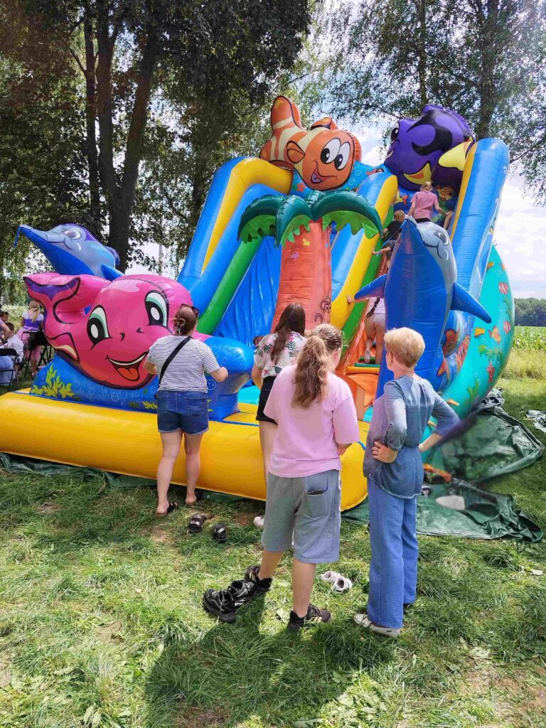 Na zdjęciu widoczna jest kolorowa, dmuchana zjeżdżalnia w kształcie zamku, ozdobiona motywami podwodnego świata — rybami, delfinami i koralowcami. Na zjeżdżalni bawią się dzieci, wspinając się na jej szczyt, podczas gdy dorośli stoją wokół, obserwując zabawę. W tle znajdują się drzewa i zielona trawa. Na ziemi widoczne są porzucone buty, które dzieci zdjęły przed wejściem na zjeżdżalnię, co dodaje scenie naturalności i beztroski. Atmosfera zdjęcia jest pełna radości, zabawy i rodzinnego wypoczynku.