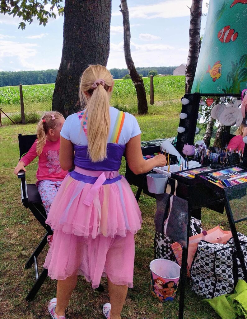 Na zdjęciu widoczne są dwie osoby podczas malowania twarzy na świeżym powietrzu. Dorosła kobieta stoi tyłem do aparatu, ubrana w kolorową sukienkę z elementami różu, fioletu i żółci, a jej długie blond włosy są związane w kucyk. Przed nią siedzi dziecko ubrane w różową bluzkę i wzorzyste spodnie w kwiaty. Na stole między nimi znajdują się przybory do malowania: pędzle, farby i lusterko. W tle widoczne są drzewa, ogrodzenie i otwarta przestrzeń.