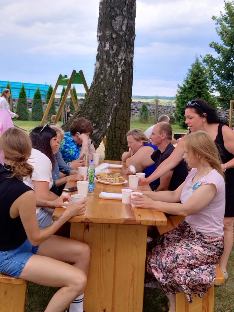 Zdjęcie ukazuje grupę osób siedzących przy drewnianym stole na świeżym powietrzu, otoczonym trawą, drzewami i ogrodzeniem. Na stole znajdują się talerze, kubki i butelka z napojem, co sugeruje, że uczestnicy wspólnie jedzą i rozmawiają. W tle można dostrzec huśtawkę, dodającą rodzinnego charakteru scenie. Atmosfera wydaje się swobodna i towarzyska.