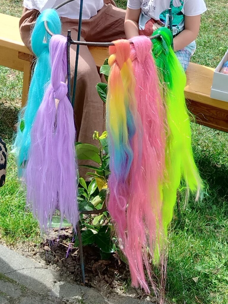 Zdjęcie przedstawia metalowy stojak, na którym zawieszone są kolorowe sztuczne włosy — w odcieniach fioletu, niebieskiego, różu, zieleni oraz tęczy. W tle, na drewnianej ławce siedzą dwie osoby — jedna z nich to dziecko. Scena ma charakter plenerowy: widać trawę i naturalne światło.