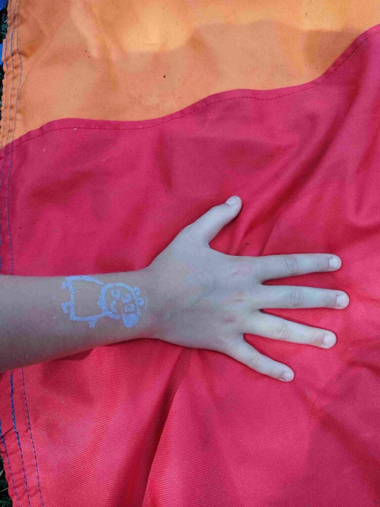 Na zdjęciu widoczna jest ręka dziecka spoczywająca na materiale w kolorze czerwonym. Na nadgarstku znajduje się niebieski tatuaż zmywalny przedstawiający kreskówkową postać, co dodaje zdjęciu dziecięcego uroku i beztroski. Cała scena wydaje się mieć charakter kreatywnej zabawy.