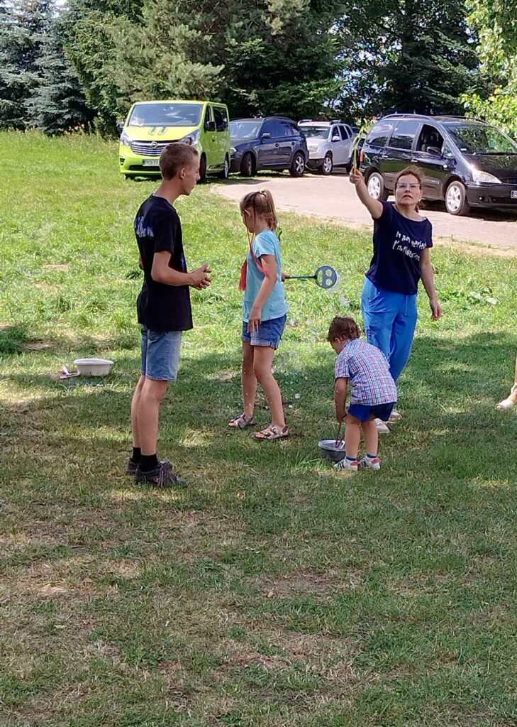 Na zdjęciu widoczna jest grupa dzieci bawiących się na zielonej trawie podczas plenerowego wydarzenia. Jedno dziecko trzyma rakietkę do badmintona, inne pochyla się nad miską z wodą, co może sugerować zabawy sensoryczne lub tematyczne. W tle znajdują się zaparkowane samochody oraz drzewa, które tworzą naturalne otoczenie. Dzieci mają na sobie letnie ubrania, a cała scena oddaje klimat aktywnej zabawy, integracji i radości spędzania czasu na świeżym powietrzu.