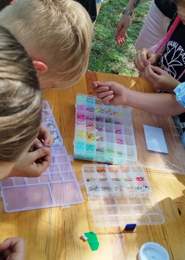 Na zdjęciu widoczna jest grupa dzieci siedzących przy drewnianym stole, skupionych na tworzeniu biżuterii z kolorowych koralików. Na stole rozłożone są plastikowe organizery z przegródkami, w których znajdują się różnorodne koraliki — od małych, jednokolorowych po ozdobne z oczkami i literami. Dzieci nawlekają koraliki na nitki, układając własne wzory. Widać tylko ich ręce, co podkreśla zaangażowanie i kreatywność w pracy twórczej. Cała scena emanuje spokojem i koncentracją.