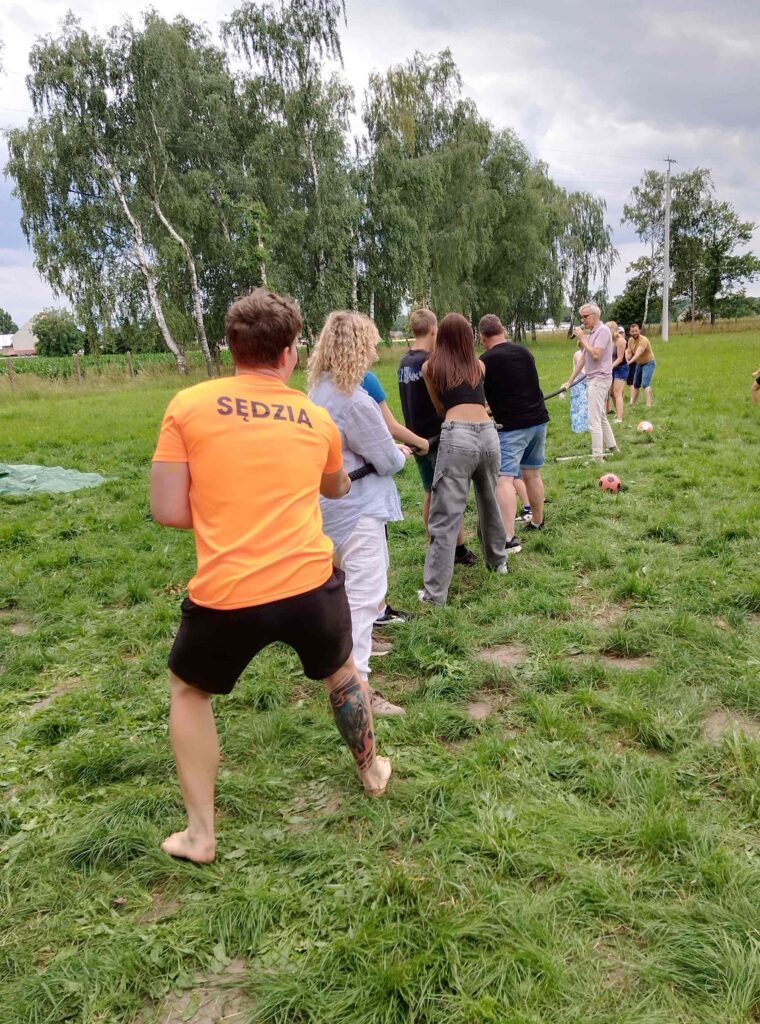 Zdjęcie przedstawia grupę osób uczestniczących w zabawie przeciągania liny na świeżym powietrzu. Centralną postacią na pierwszym planie jest osoba w pomarańczowej koszulce z napisem „SĘDZIA”. Po obu stronach liny stoją zawodnicy — dzieci i dorośli — napinając ją z pełnym zaangażowaniem. W tle widać zieloną trawę, drzewa oraz dwie piłki leżące na ziemi, co może sugerować inne aktywności sportowe odbywające się równolegle. Atmosfera jest energiczna i integracyjna.