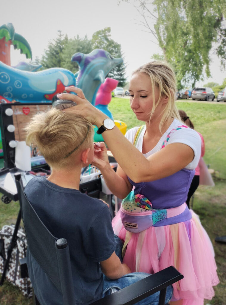 Na zdjęciu widoczne jest dziecko siedzące na krześle, któremu osoba w kolorowym stroju maluje twarz. Osoba ta ma na sobie różową spódnicę i fioletowy top. W tle znajdują się dmuchane zabawki — w tym delfin i palma — oraz zaparkowane samochody, co wskazuje, że scena odbywa się na świeżym powietrzu