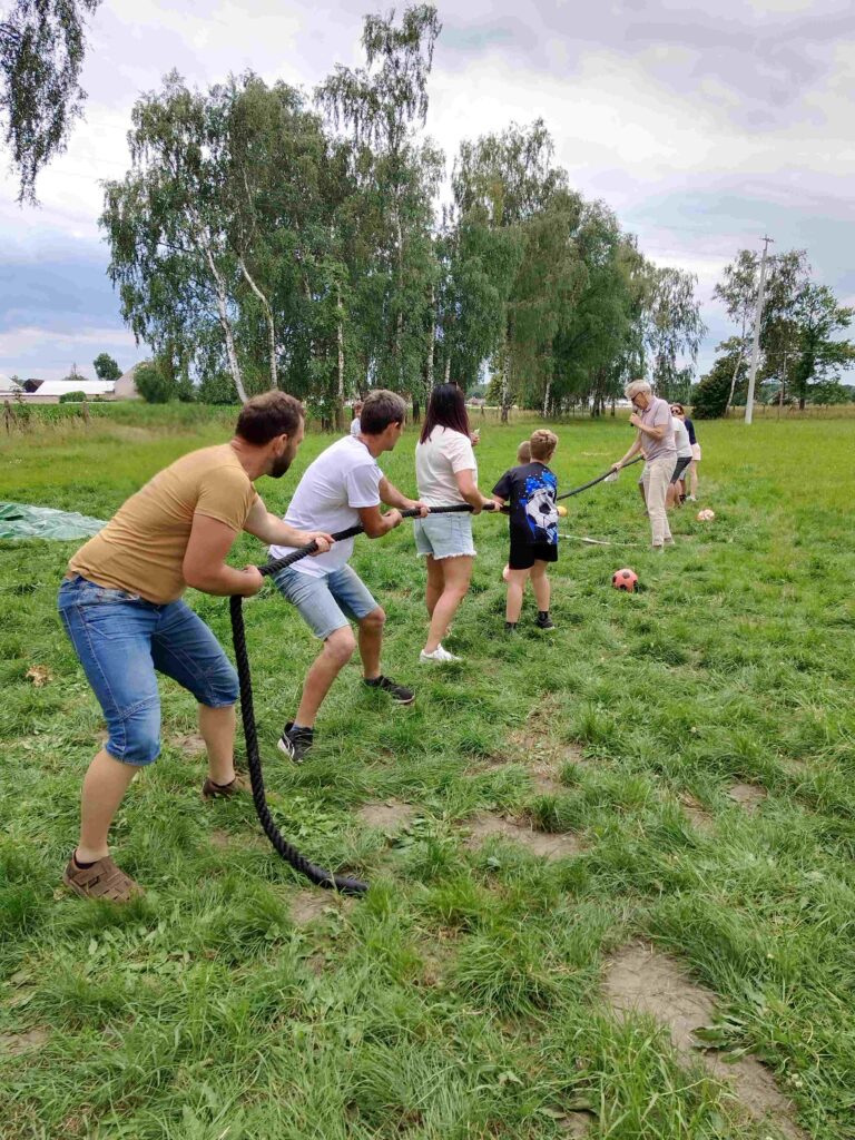Na zdjęciu przedstawiona jest grupa osób uczestniczących w zabawie przeciągania liny na świeżym powietrzu. Na pierwszym planie cztery osoby, w tym dzieci i dorośli, mocno trzymają linę, napinając ją w kierunku przeciwnym do dwóch osób po drugiej stronie. W tle widoczna jest zielona trawa, drzewa oraz fragmenty budynków, co sugeruje, że wydarzenie odbywa się na otwartej przestrzeni. Uczestnicy wyglądają na zaangażowanych i skupionych, co oddaje energię oraz radość wspólnej zabawy i integracji.