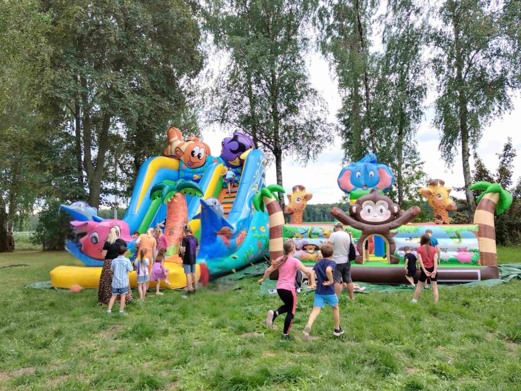 Zdjęcie przedstawia grupę dzieci bawiących się na kolorowym, dmuchanym placu zabaw z motywami zwierząt. Centralnym elementem jest duży, dmuchany zamek ozdobiony grafikami przedstawiającymi m.in. żyrafę, słonia, małpę, rybę i delfina. Dzieci wspinają się na konstrukcję i korzystają ze zjeżdżalni, otoczone naturą — w tle widoczne są drzewa. Scena oddaje radosną i beztroską atmosferę zabawy na świeżym powietrzu.