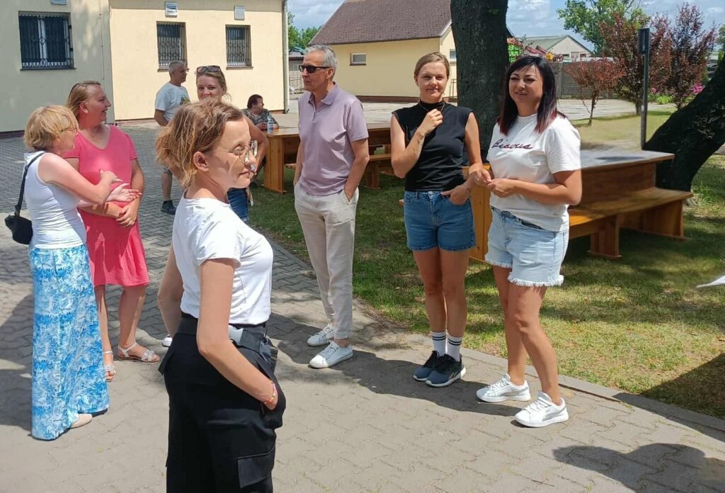 Na zdjęciu widać grupę siedmiu osób stojących na utwardzonym placu na świeżym powietrzu, w pobliżu trawnika i budynku o jasnobeżowej elewacji z oknami zabezpieczonymi kratami. Część osób prowadzi rozmowę, a ich stroje — koszulki, szorty, sandały — wskazują na ciepły letni dzień. W tle znajduje się drewniana konstrukcja przypominająca ławkę lub stół oraz zieleń w postaci krzewów i drzew. Ogólna atmosfera zdjęcia jest swobodna, towarzyska i letnia.