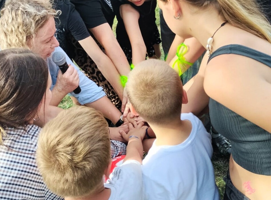 Na zdjęciu widoczna jest grupa osób stojących w kręgu, które wspólnie układają dłonie w centralnym stosie — gest jedności i współpracy. W centrum znajdują się dwaj chłopcy, którzy również dołączają swoje ręce. Jedna z osób trzyma mikrofon, co sugeruje prowadzenie zabawy lub ogłoszenie. Uczestnicy ubrani są w codzienne stroje, a niektórzy mają ozdobne bransoletki. Tło wskazuje na otwartą przestrzeń. Scena oddaje ducha wspólnoty, zaangażowania i pozytywnej energii.