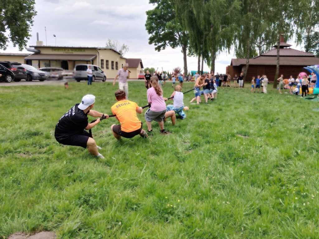 Zdjęcie przedstawia grupę osób biorących udział w przeciąganiu liny na trawie podczas wydarzenia plenerowego. Na pierwszym planie widocznych jest dwóch mężczyzn ubranych w koszulki z napisem „SĘDZIA”. W tle dostrzec można budynki, zaparkowane samochody oraz innych uczestników i obserwatorów zabawy. Atmosfera zdjęcia jest pełna energii i wspólnoty.