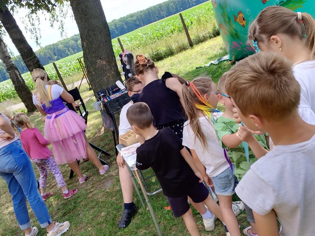 Zdjęcie przedstawia grupę dzieci na świeżym powietrzu, stojących w kolejce do stanowiska animacyjnego. Na pierwszym planie znajduje się osoba ubrana w różową tiulową spódnicę i fioletowy top — animatorka — która wykonuje malowanie twarzy lub inne atrakcje dla dzieci. W tle widać drzewa, pole kukurydzy oraz duży, kolorowy balon dekoracyjny z motywem ryb i morskich stworzeń. Dzieci mają różnorodne stroje — część z nich ma pomalowane twarze albo warkoczyki w kolorowych pasemkach. Cała scena emanuje radością, beztroską i atmosferą festynu rodzinnego.