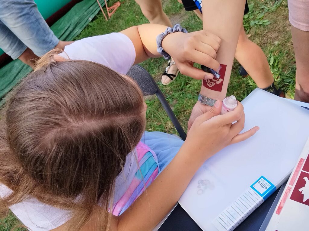Na zdjęciu widoczne jest dziecko wykonujące tymczasowy tatuaż na przedramieniu drugiej osoby. Malowanie odbywa się za pomocą szablonu przyklejonego do skóry i aplikatora z farbą, co sugeruje skupienie i kreatywność twórcy. Obok znajduje się biały pojemnik z napisem „Office” oraz różne drobne przedmioty, prawdopodobnie potrzebne do wykonania tatuażu. Całość dzieje się na świeżym powietrzu a scena oddaje klimat zabawy, integracji i twórczej aktywności — typowej dla festynu, warsztatów lub rodzinnej imprezy.