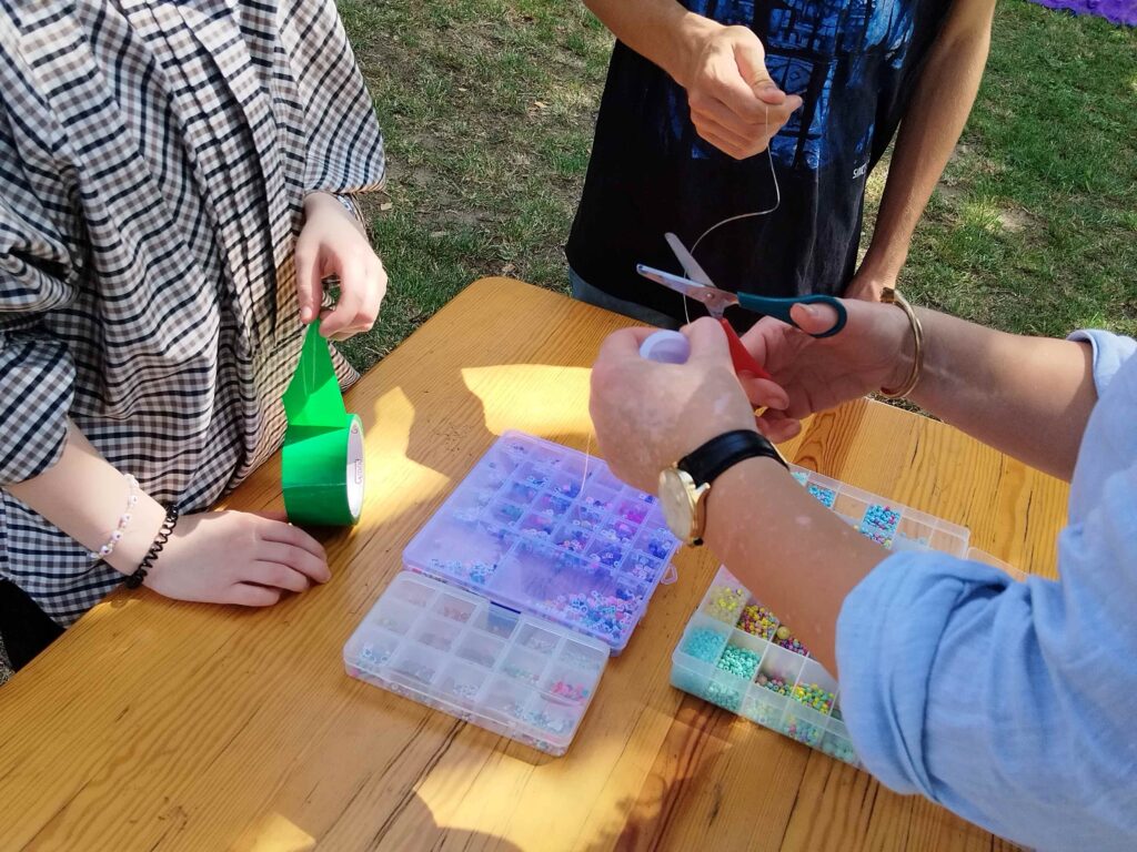 Zdjęcie przedstawia trzy osoby zaangażowane w twórczą pracę przy stole na świeżym powietrzu. Jedna osoba trzyma zieloną taśmę klejącą, druga przecina materiał przy pomocy nożyczek, a trzecia operuje cienkim drutem. Na stole rozłożone są dwa pudełka z kolorowymi koralikami, co sugeruje, że grupa bierze udział w rękodzielniczych warsztatach — być może tworzą biżuterię, ozdoby lub dekoracje. Tło wskazuje na plenerowe otoczenie, dodając scenie lekką i kreatywną atmosferę.