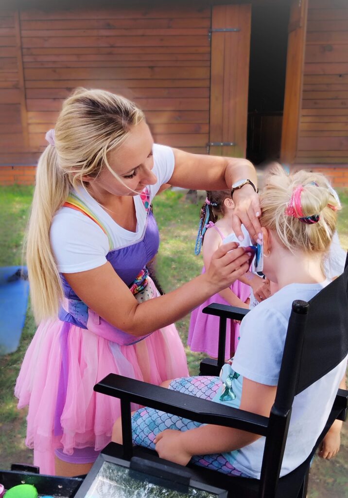 Na zdjęciu widoczne jest dziecko siedzące na krześle, którego twarz jest malowana przez osobę przebraną w barwny kostium — przypominający baletnicę, z różową tiulową spódnicą. Dziecko ma na sobie niebieski strój w łuskowaty wzór, przypominający syreni ogon. W tle znajduje się drewniany budynek, a wokół widać inne dzieci ubrane w kolorowe stroje, co sugeruje, że wydarzenie ma charakter zabawowy — piknik rodzinny.