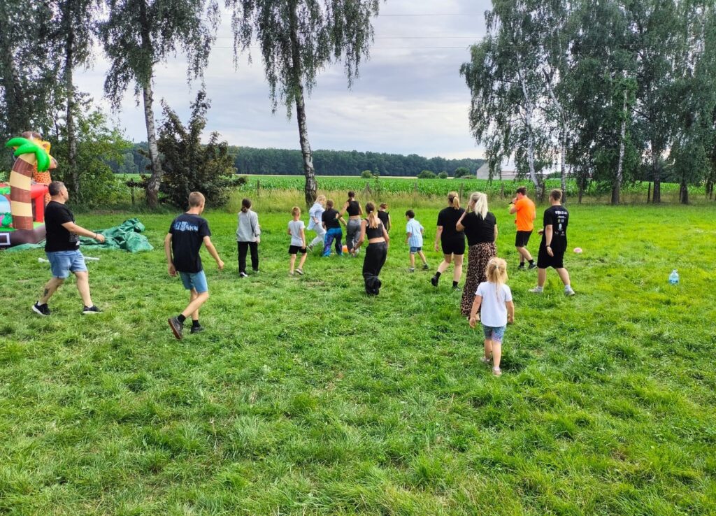 Zdjęcie przedstawia grupę dzieci i dorosłych bawiących się na zielonej trawie podczas plenerowego wydarzenia. W tle widoczne są drzewa oraz pole uprawne, co dodaje naturalnego uroku scenerii. Po lewej stronie znajduje się kolorowy dmuchany zamek, obok którego leży zielona plandeka — mogą to być elementy placu zabaw lub strefy animacyjnej. Uczestnicy ubrani są swobodnie: dzieci w krótkich spodenkach, dorośli w koszulkach i bluzach, co wskazuje na letni dzień i nieformalny charakter spotkania. Scena emanuje radością, ruchem i wspólnotową atmosferą, typową dla festynów rodzinnych i imprez plenerowych.