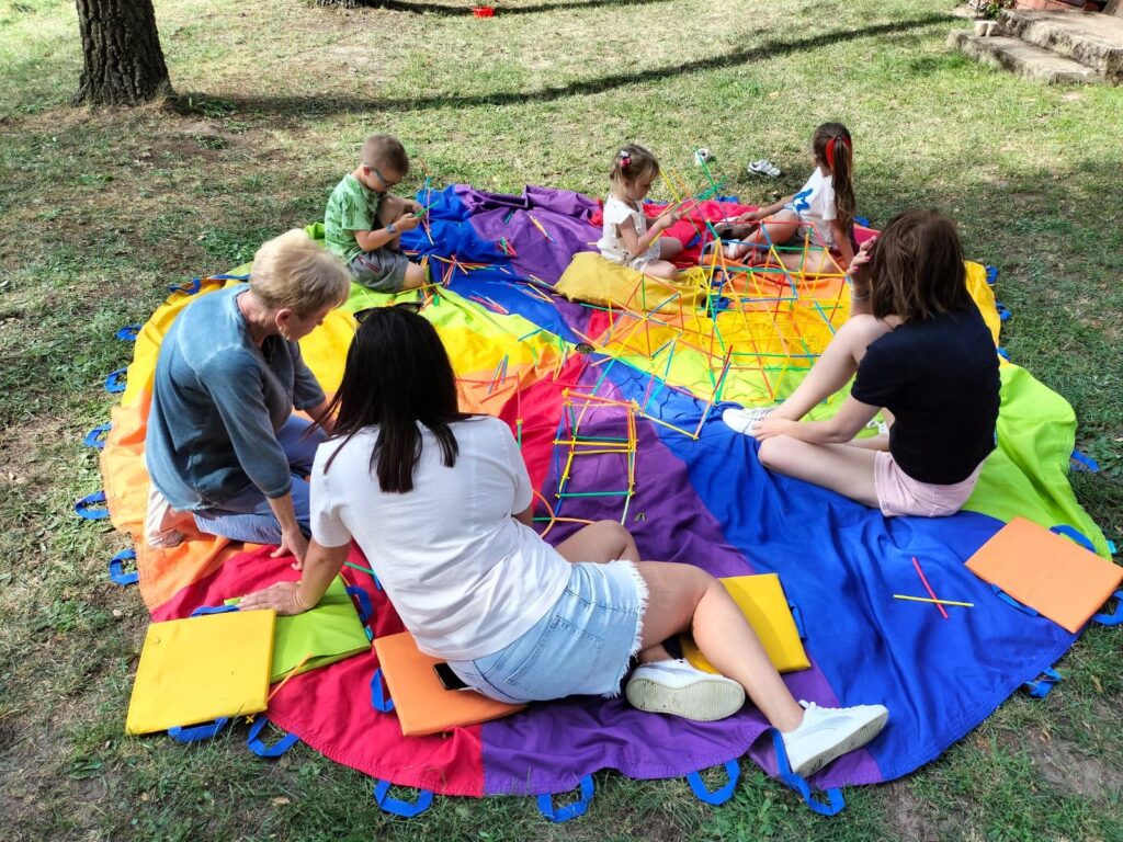 Na zdjęciu widoczna jest grupa dzieci i dorosłych siedzących na wielobarwnej macie ułożonej na trawie podczas wydarzenia plenerowego. Uczestnicy zabawy zajmują się konstruowaniem różnych form z kolorowych patyczków, rozwijając kreatywność i zdolności manualne. Wokół maty rozmieszczono kolorowe poduszki, na których siedzą uczestnicy — dominują barwy żółta, czerwona, niebieska, zielona i fioletowa. W tle widać naturalne otoczenie z drzewami, co podkreśla atmosferę zabawy na świeżym powietrzu.