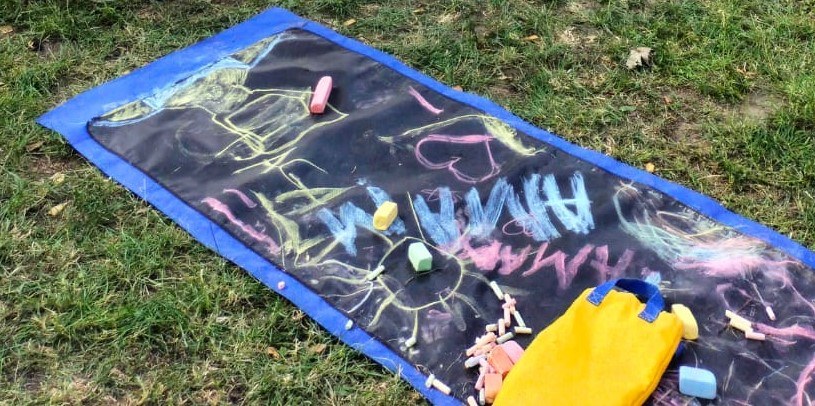 Na zdjęciu znajduje się mata do rysowania kredą rozłożona na zielonej trawie. Na powierzchni maty widać kolorowe rysunki wykonane kredą — między innymi serce, ludzką sylwetkę oraz napis „HUMAN”. Obok maty znajduje się torba z kredami w różnych kolorach. Całość ukazuje moment twórczej aktywności na świeżym powietrzu.