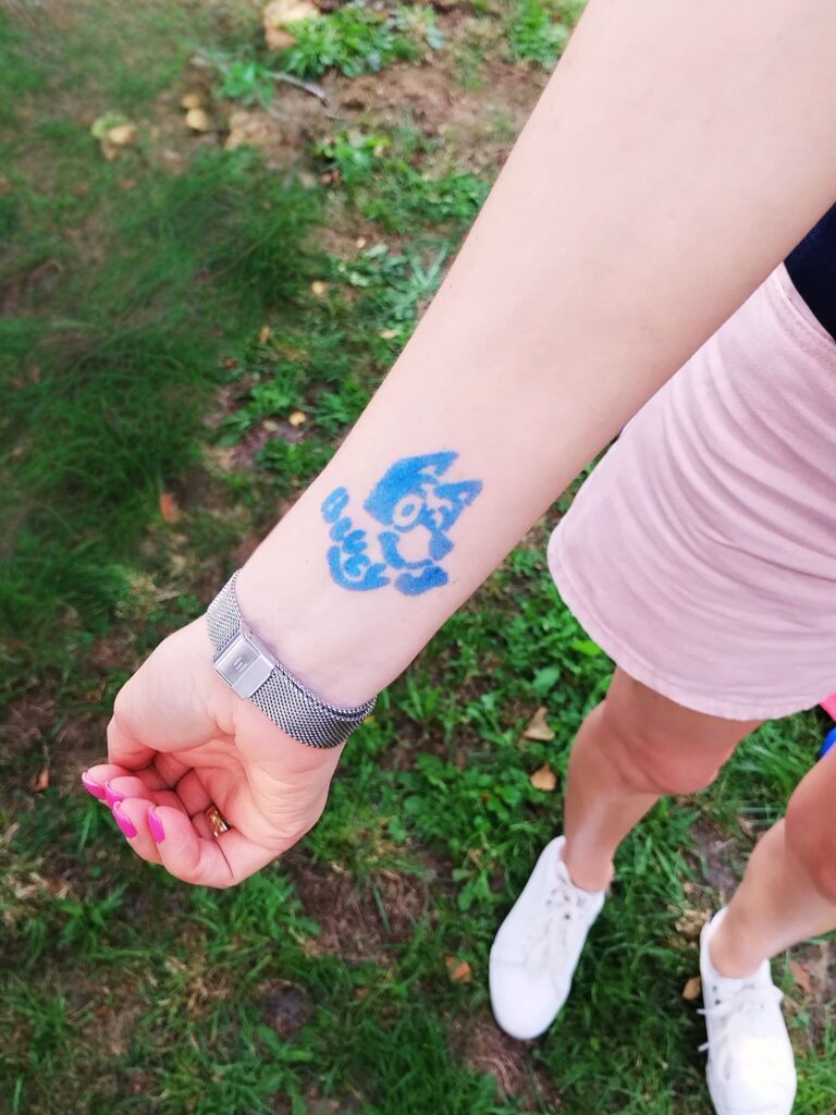 Zdjęcie przedstawia przedramię osoby z wyrazistym tatuażem w odcieniach niebieskiego. Tatuaż przedstawia stylizowaną głowę wilka, a pod nią widnieje napis (którego treść nie została podana). Osoba ubrana jest w różowe szorty, białe buty sportowe i nosi srebrny zegarek na nadgarstku. Tło zdjęcia to trawiasta powierzchnia i ziemia, co sugeruje, że fotografia została wykonana na świeżym powietrzu.