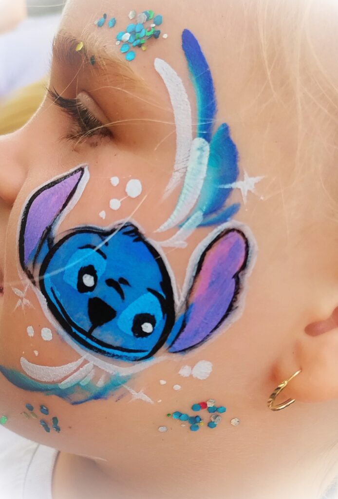 Zbliżenie na policzek i ucho osoby, której twarz została pomalowana w artystyczny sposób. Malunek składa się z falujących linii w odcieniach niebieskiego, zielonego i białego, a także drobnych kolorowych kropek — m.in. niebieskich, zielonych i czerwonych. Osoba nosi małą, złotą kolczykową obręcz. Stylizacja twarzy wskazuje na udział w imprezie z animacjami i malowaniem twarzy.