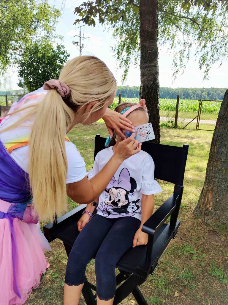 Na zdjęciu przedstawiono dziecko siedzące na krześle reżyserskim na świeżym powietrzu, w otoczeniu drzew i ogrodzenia. Dziecko ma na sobie białą koszulkę z nadrukiem Minnie Mouse, czarne legginsy oraz opaskę z uszami na głowie. Osoba malująca twarz dziecka ma długie blond włosy związane w kucyk, kolorowy strój i używa szablonu z rysunkiem psa do nałożenia niebieskiej farby na twarz dziecka. Scena oddaje atmosferę zabawy i kreatywnej aktywności.