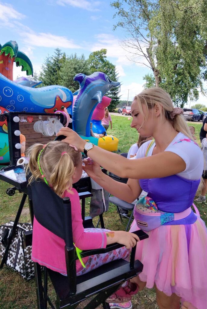 Zdjęcie przedstawia scenę z festynu plenerowego, gdzie dziecko siedzi na krześle, a kobieta w barwnym stroju maluje jego twarz. Kobieta ubrana jest w kolorową tiulową spódnicę typu tutu oraz fioletowy gorset, co nadaje jej wygląd animatorki zabaw. W tle widoczne są dmuchane dekoracje — delfiny i inne morskie stwory — które wzmacniają wakacyjną, radosną atmosferę wydarzenia. Scena emanuje kreatywnością, dziecięcą radością i rodzinno-rekreacyjnym klimatem.