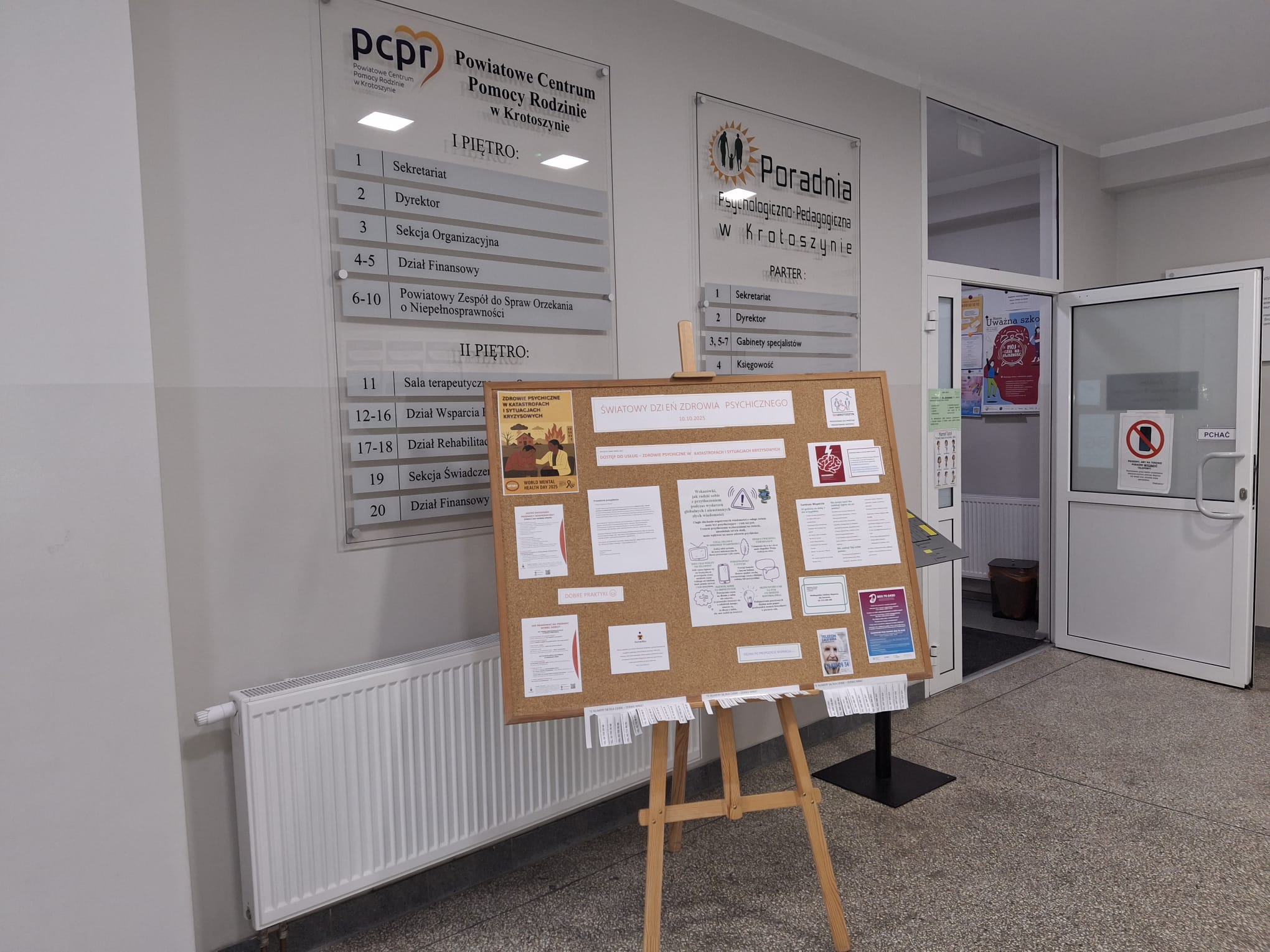 Wnętrze budynku Powiatowego Centrum Pomocy Rodzinie i Poradni Psychologiczno-Pedagogicznej w Krotoszynie. Na ścianach znajdują się tablice informacyjne z nazwami działów i numerami pokoi. Na środku stoi tablica korkowa na sztaludze z ogłoszeniami i plakatami, m.in. dotyczącymi Światowego Dnia Zdrowia Psychicznego. Po prawej stronie widać otwarte drzwi prowadzące do dalszej części budynku, z widocznymi plakatami informacyjnymi na ścianach.