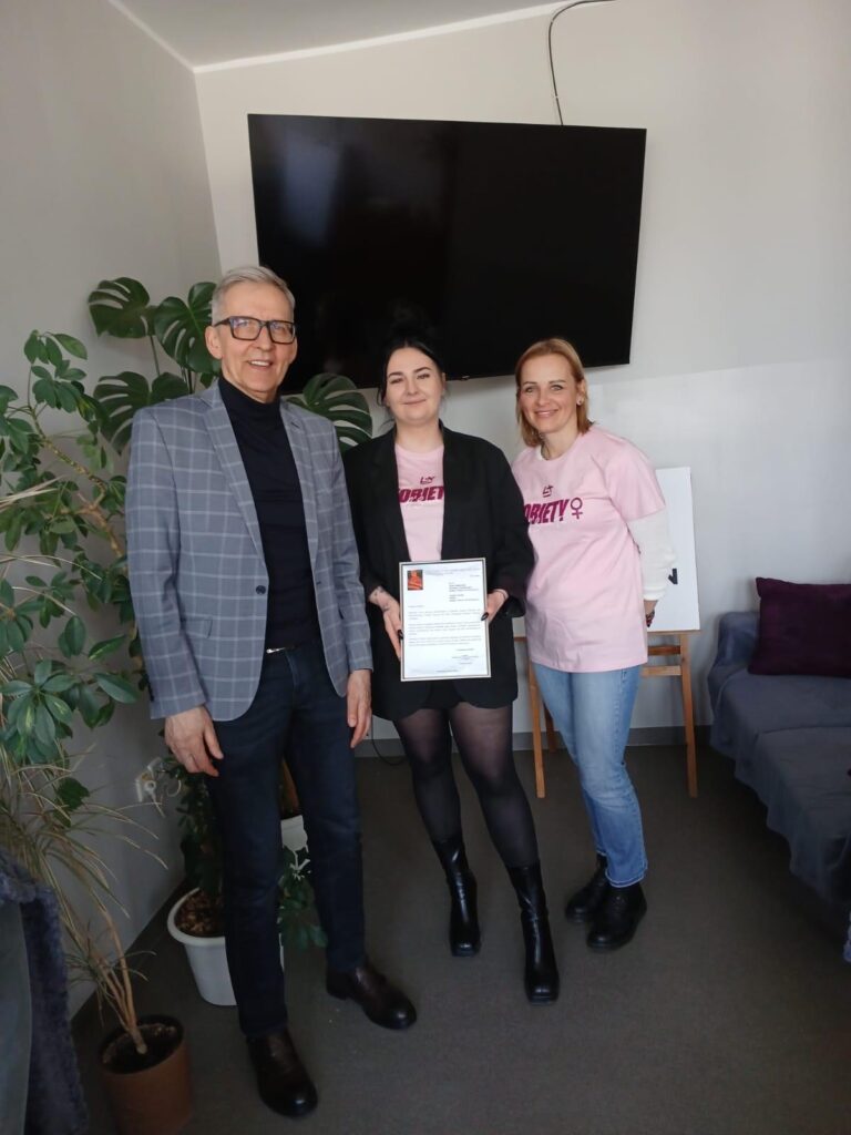 Zdjęcie pamiątkowe z wręczenia dokumentu. Po lewej mężczyzna w okularach i kraciastej marynarce. W środku kobieta w czarnej marynarce trzymająca certyfikat. Po prawej kobieta w różowym t-shircie. W tle widać roślinę doniczkową (monstera) oraz duży ekran telewizora.