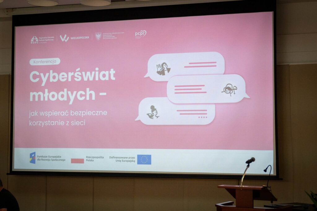 Ekran projekcyjny z tytułowym slajdem konferencji „Cyberświat młodych – jak wspierać bezpieczne korzystanie z sieci". Różowe tło z grafikami przedstawiającymi dymki czatu z sylwetkami dzieci. Widoczne logotypy organizatorów: ROPS w Poznaniu, Wielkopolska, jednostka samorządu województwa wielkopolskiego oraz PCPR. Na dole slajdu logotypy Funduszy Europejskich dla Rozwoju Społecznego, flaga Polski i Unii Europejskiej.
