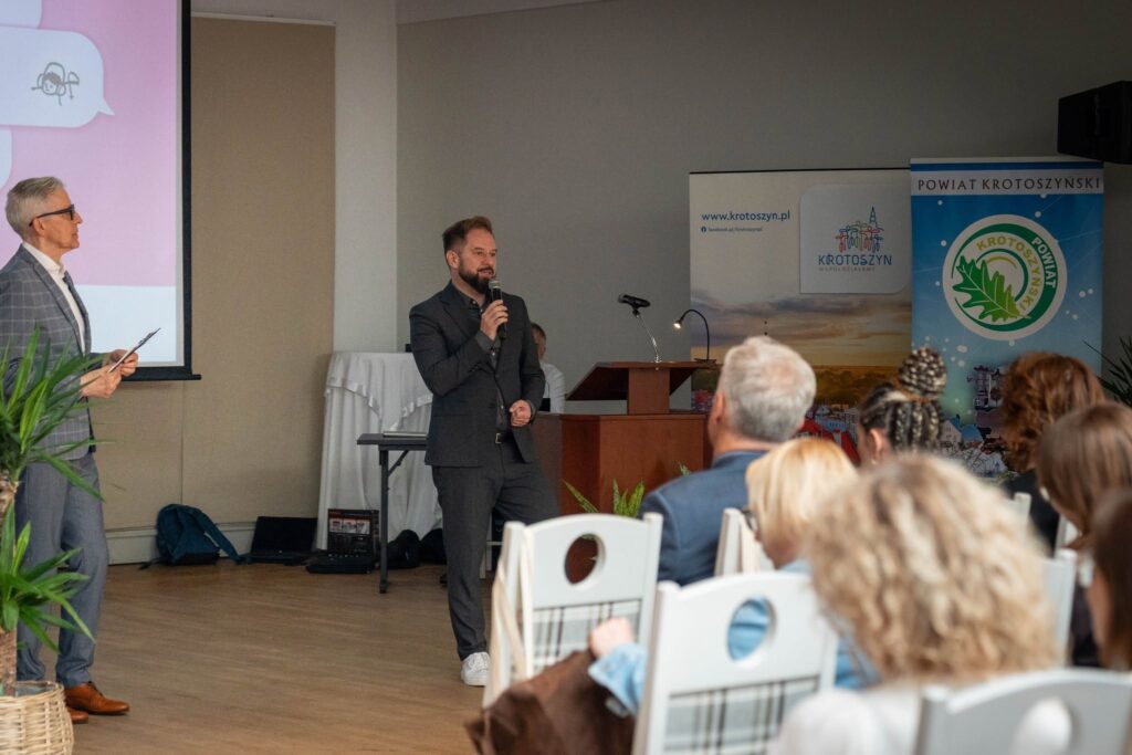 Dwóch mężczyzn na sali konferencyjnej. Jeden stoi z mikrofonem i przemawia do zgromadzonych uczestników, drugi trzyma notes i przysłuchuje się. W tle widoczny ekran z fragmentem prezentacji oraz roll-upy Powiatu Krotoszyńskiego i miasta Krotoszyn. Na pierwszym planie siedzący tyłem uczestnicy konferencji.