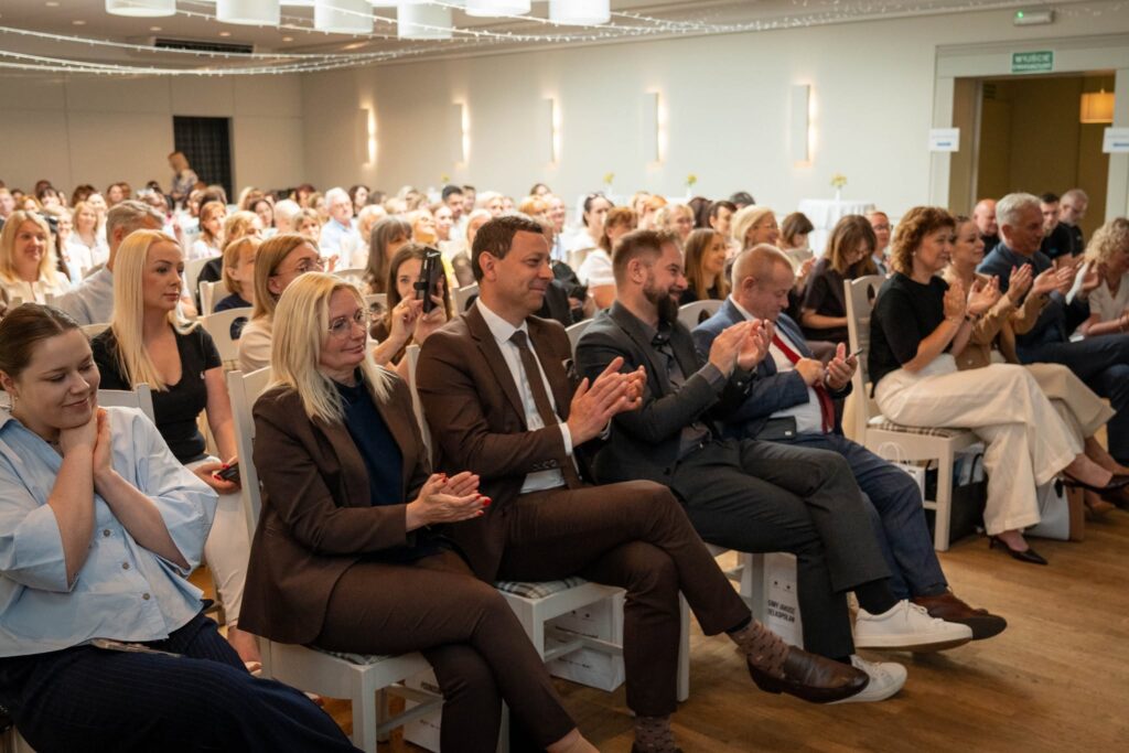 Wypełniona sala konferencyjna – widok na kilkadziesiąt osób siedzących na białych krzesłach. Uczestnicy w pierwszych rzędach klaszczą. Na pierwszym planie widoczna kobieta w brązowym stroju i mężczyzna w brązowym garniturze. Sala jest jasna, z dekoracyjnymi lampami na ścianach.