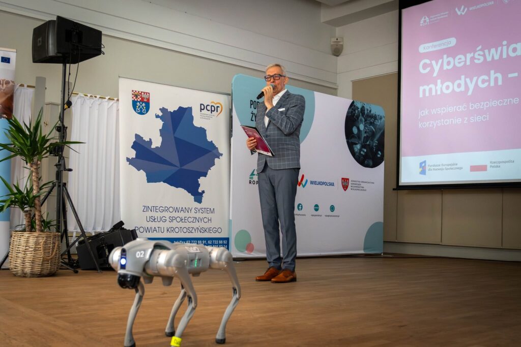 Mężczyzna w szarej marynarce w kratę przemawia z mikrofonem na sali konferencyjnej. Na podłodze przed nim stoi czworonożny robot (pies robotyczny). W tle roll-up PCPR Krotoszyn z mapą powiatu i napisem „Zintegrowany System Usług Społecznych Powiatu Krotoszyńskiego" oraz ekran z tytułem konferencji „Cyberświat młodych".