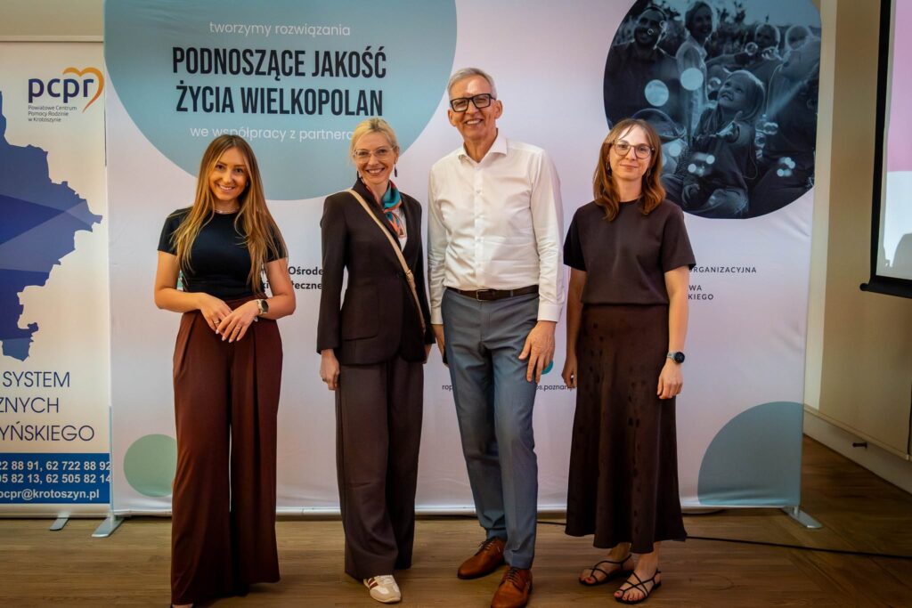 Cztery osoby pozują do zdjęcia grupowego na tle roll-upu z hasłem „Podnoszące jakość życia Wielkopolan". Od lewej: młoda kobieta w czarnej bluzce i brązowych spodniach, kobieta w żakiecie z apaszką, wysoki mężczyzna w białej koszuli oraz kobieta w brązowej sukience i okularach.
