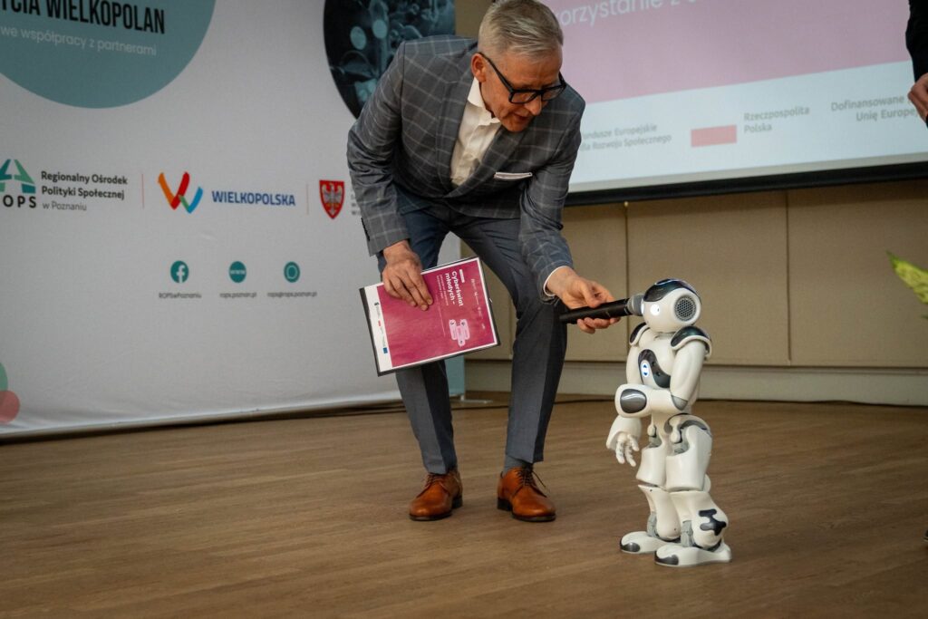 Mężczyzna w szarej marynarce w kratę pochyla się i podaje mikrofon małemu humanoidalnemu robotowi (NAO) stojącemu na podłodze sali konferencyjnej. W tle baner z logotypami ROPS w Poznaniu, Wielkopolska i samorządu województwa oraz ekran z prezentacją konferencji.